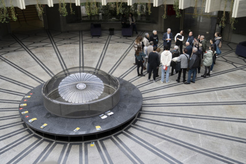 Vista cenital de un espacio interior amplio con suelo de patrón radial. En el centro hay una estructura circular acristalada rodeada de folletos, y a un lado un grupo de personas escucha a un guía, lo que sugiere una visita o actividad cultural en un edificio público.